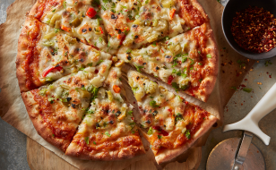 Cut pizza with various toppings on top of parchment paper and wooden cutting board with side pizza cutter and bowl of red pepper flakes.