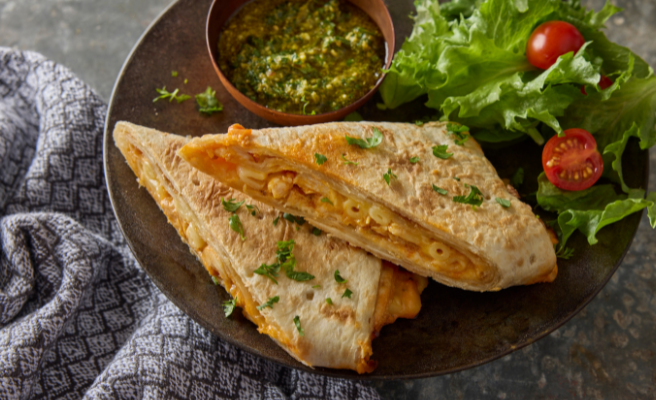 Two mac n’ cheese wraps stacked on a plate with a small side salad and small bowl of cilantro chutney sauce.