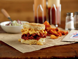 Pulled Pork Mac ‘n Cheese Biscuit Sandwich recipe on brown paper with chips, coleslaw, and condiments in background.