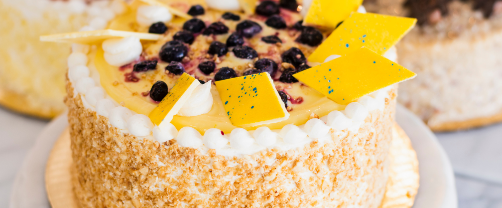 A vibrant, artisan layer cake topped with citrus glaze, blueberries, and decorative chocolate shards, showcasing the craftsmanship and creativity of Crème de la Cocoa desserts.