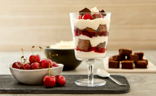 A parfait glass filled with layers of chopped brownies and whipped cream next to a bowl of cherries on a serving platter