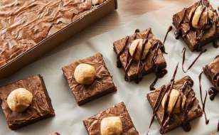 Cut brownie squares in front of pan of baked brownies with bowl of melted chocolate in background.