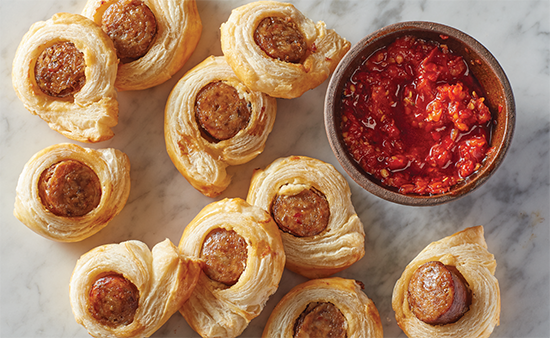 Pigs in a blanket wrapped with biscuits on a granite counter with chili dipping sauce on the side.